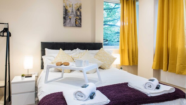 Habitación de hotel con una cama, una mesita de noche con lámpara, una ventana con cortinas amarillas y un cuadro en la pared.