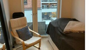 Ein kleiner, moderner Raum mit einem Bett, einem Sessel und Blick auf ein Gebäude durch eine Glastür.