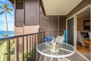 A balcony with a table and chairs, a view of the ocean, and a door leading to a room with a bed and a window.