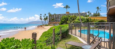 A poolside area with a view of the beach and ocean, surrounded by greenery and a building with multiple floors.