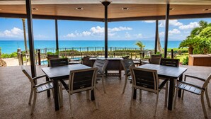 A covered outdoor seating area with tables and chairs, overlooking a pool and the ocean.