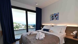 A hotel room with a large bed, a bedside table with a lamp, and a view of the city through a large window.