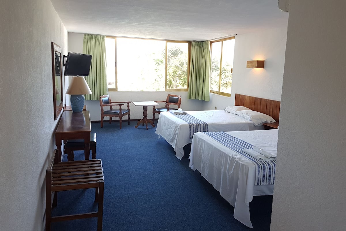 Habitación de hotel con dos camas, un escritorio con lámpara, una silla y una mesita.