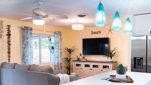 A modern living room with a sofa, a dining area with a kitchen island, a ceiling fan, and a TV mounted on the wall.