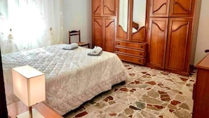 A bedroom with a bed, a wooden wardrobe, a chair, and a nightstand with a lamp.