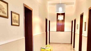 A hotel hallway with framed pictures on the walls, a wooden chair, a small round table with magazines, and a yellow table with chairs.