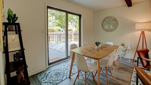 Un coin repas avec une table en bois et des chaises blanches, une bibliothèque, une lampe et, à travers une porte vitrée, une vue sur un patio extérieur.