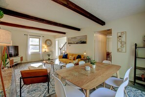A spacious living room with a wooden dining table, white chairs, a sofa with cushions, a television, and a large painting on the wall.