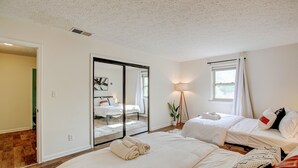 Une chambre d’hôtel avec un grand lit, un plus petit lit, un miroir, une fenêtre avec des rideaux, une lampe sur pied et une vue sur une autre chambre, visible à travers une porte coulissante.