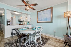 A dining area with a round table, chairs, a ceiling fan, and a kitchen in the background.