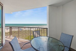A balcony with a glass table and chairs, overlooking a beach with a clear sky.