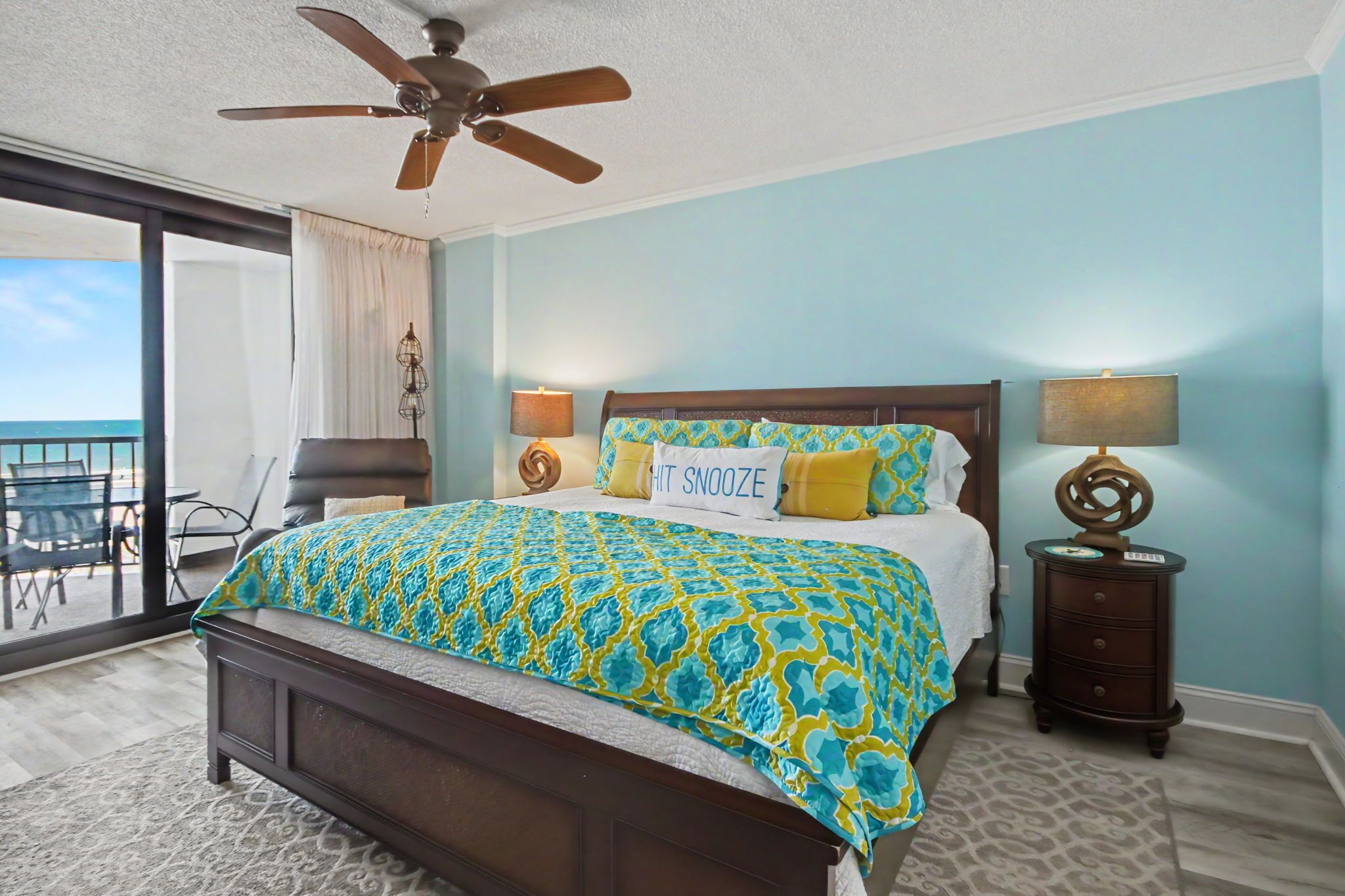 A bedroom with a bed, a ceiling fan, a nightstand, a lamp, and a view of the balcony.