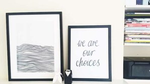 Deux œuvres d’art encadrées sont posées sur une étagère blanche : une composition abstraite en noir et blanc et une citation encadrée qui dit : « Nous sommes le produit de nos choix. »
