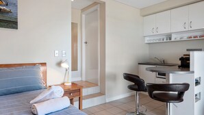 A compact hotel room with a bed, a bedside lamp, a chair, a small kitchen area with a sink and a coffee machine, and a door leading to another room.