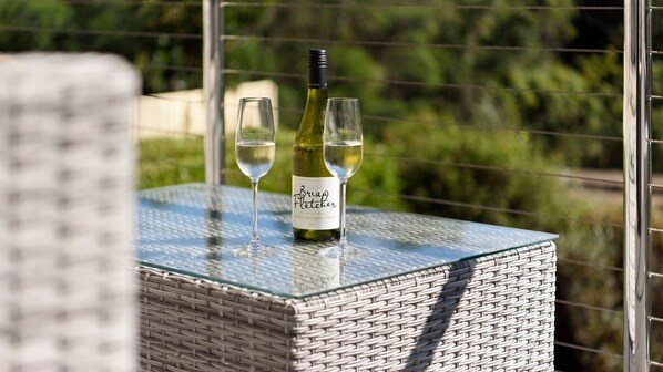 Une bouteille de Sauvignon Blanc 2021 de Brian Fletcher et deux verres posés sur une table en verre, avec une chaise en osier en arrière-plan.