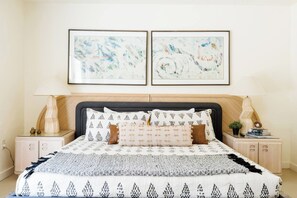 Un dormitorio con una cama, dos mesitas de noche, una mesita auxiliar, una lámpara y dos cuadros abstractos en la pared.