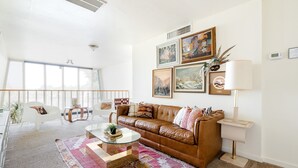 A spacious living room with a brown leather couch, a glass coffee table, and a colorful rug. There are large windows providing natural light, and the room has a modern, minimalist design.