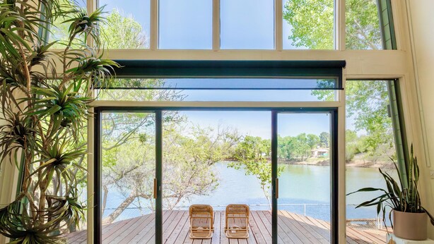 A room with a large sliding glass door leading to a deck with a view of a lake and trees. There are two wicker chairs on the deck, a wooden table, and a potted plant.