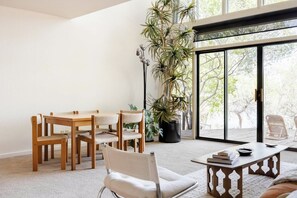 A modern living room with a wooden dining table, chairs, a large potted plant, and a view through a glass door.