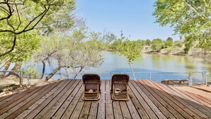 A wooden deck with two wicker chairs facing a calm lake, surrounded by trees and a fence.