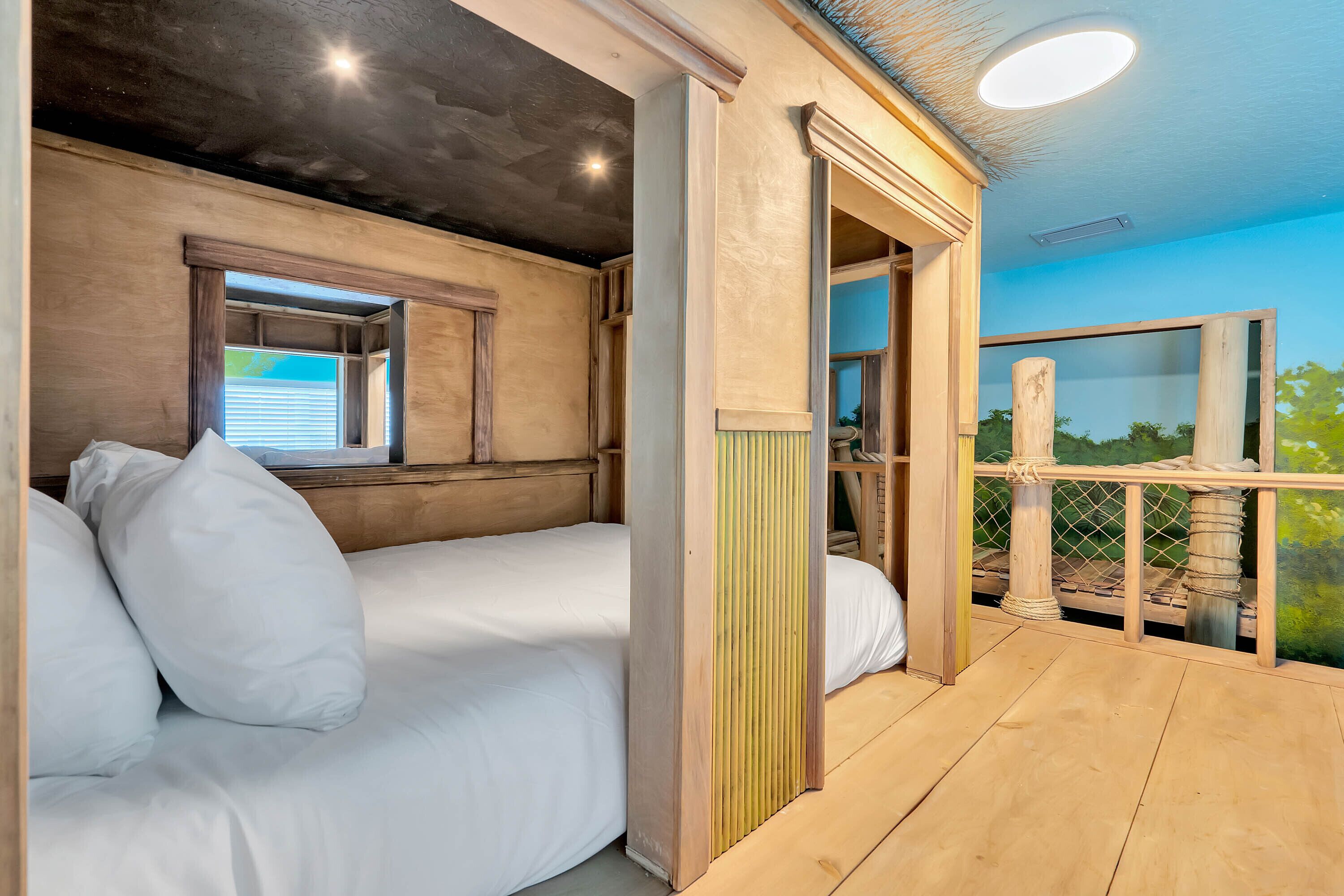 Ein Schlafzimmer mit hölzernem Kopfteil, einem Fenster mit Jalousien, einem Balkon mit Blick ins Grüne und einer Deckenleuchte.