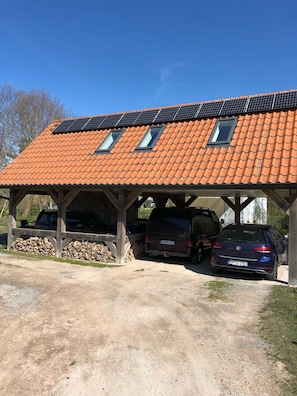 Un abri-voiture fermé avec deux voitures garées en dessous, un toit équipé de panneaux solaires et une pile de bois de chauffage sur le côté gauche.