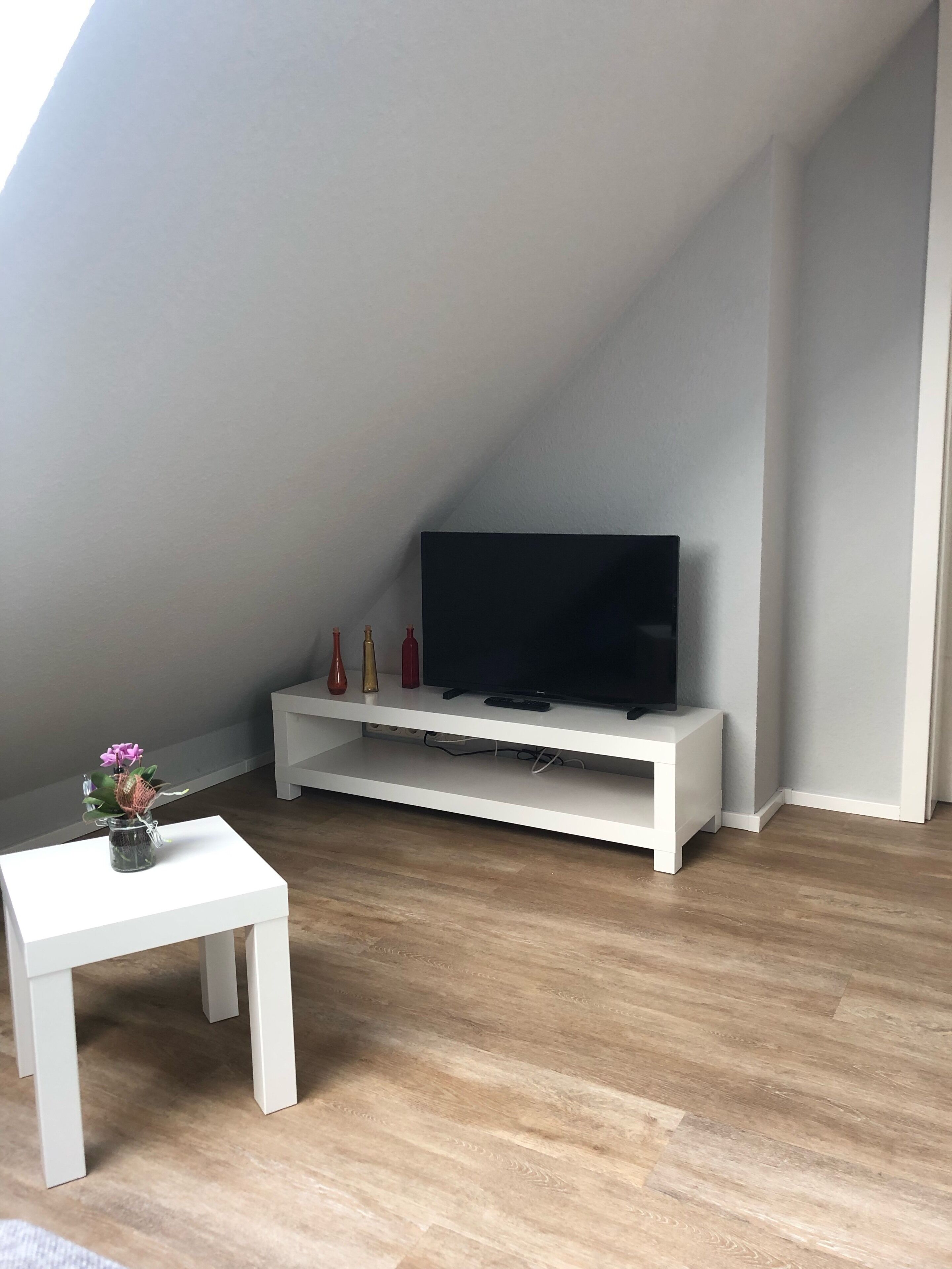 A room with a white TV stand, a flat-screen TV, a small white table with a vase of flowers, and wooden flooring.