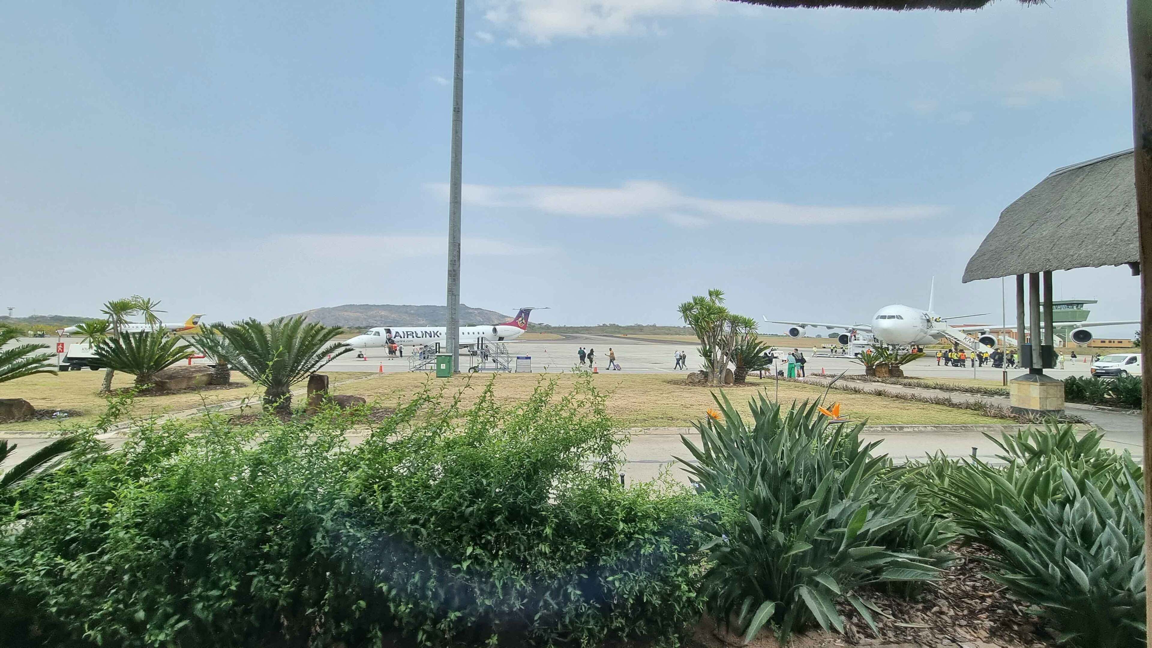 Pista de aeroporto com vários aviões, incluindo um avião comercial grande e aeronaves menores, com passageiros e equipe de solo visíveis.