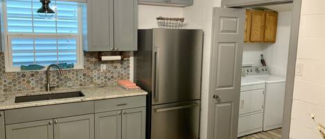 A kitchen with grey cabinets, a stainless steel refrigerator, a sink with a window above it, and a wooden door leading to a laundry area.