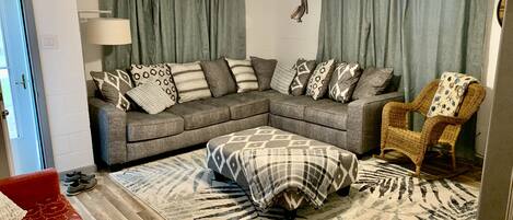 A living room with a grey sectional sofa, a patterned ottoman, a wicker chair, and a ceiling fan.