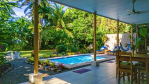 Un espacio al aire libre techado con piscina, sillas de descanso y plantas tropicales.