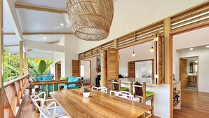 Un comedor amplio con una mesa de madera y sillas blancas, un lámpara colgante y vista a un jardín exuberante a través de ventanas grandes.