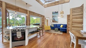 Un amplio salón con cocina, suelo de madera, un sofá azul y grandes ventanales con vistas al verdor.