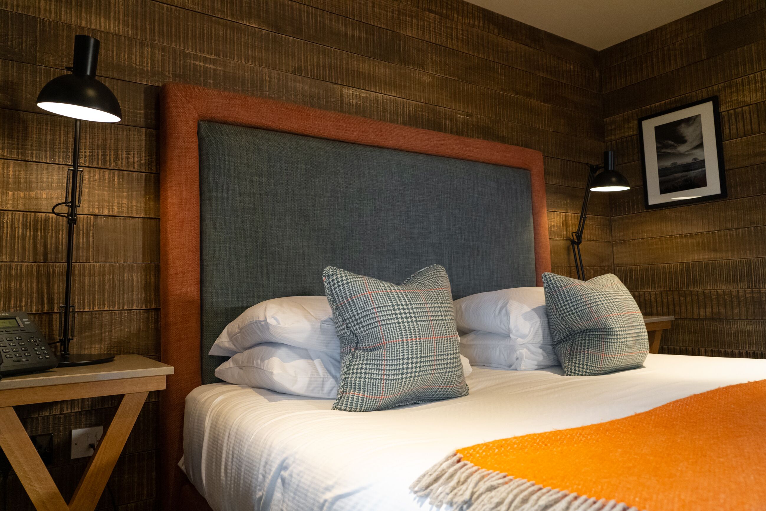 Una cama bien hecha con cabecera gris, dos almohadas y una manta naranja. Se observa una mesita de noche con una lámpara y un teléfono.