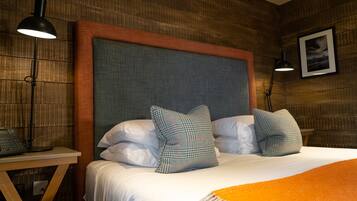 Una cama bien hecha con cabecera gris, dos almohadas y una manta naranja. Se observa una mesita de noche con una lámpara y un teléfono.
