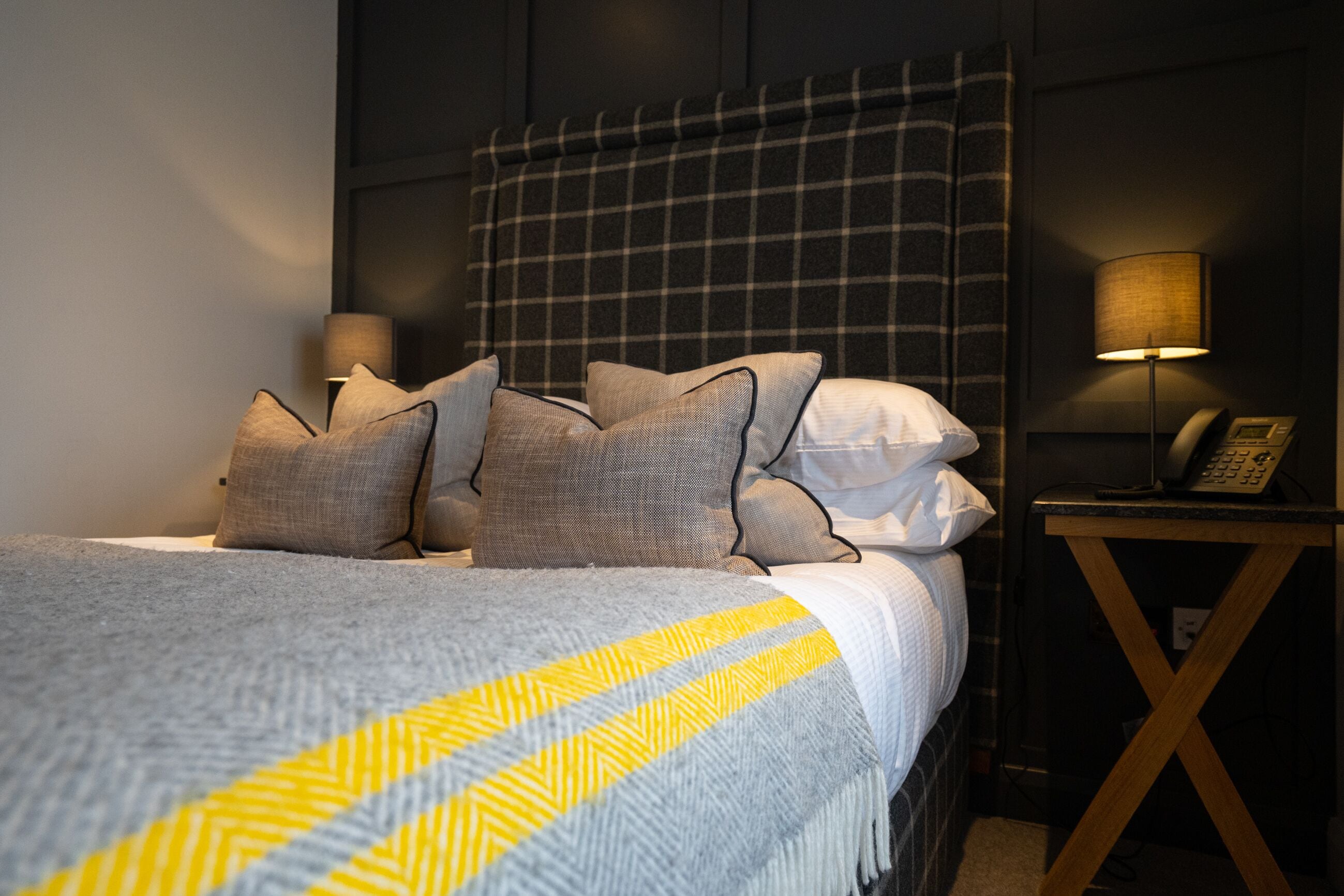 A neatly made bed with a grey and yellow striped blanket, a plaid headboard, and a bedside lamp on a wooden nightstand.
