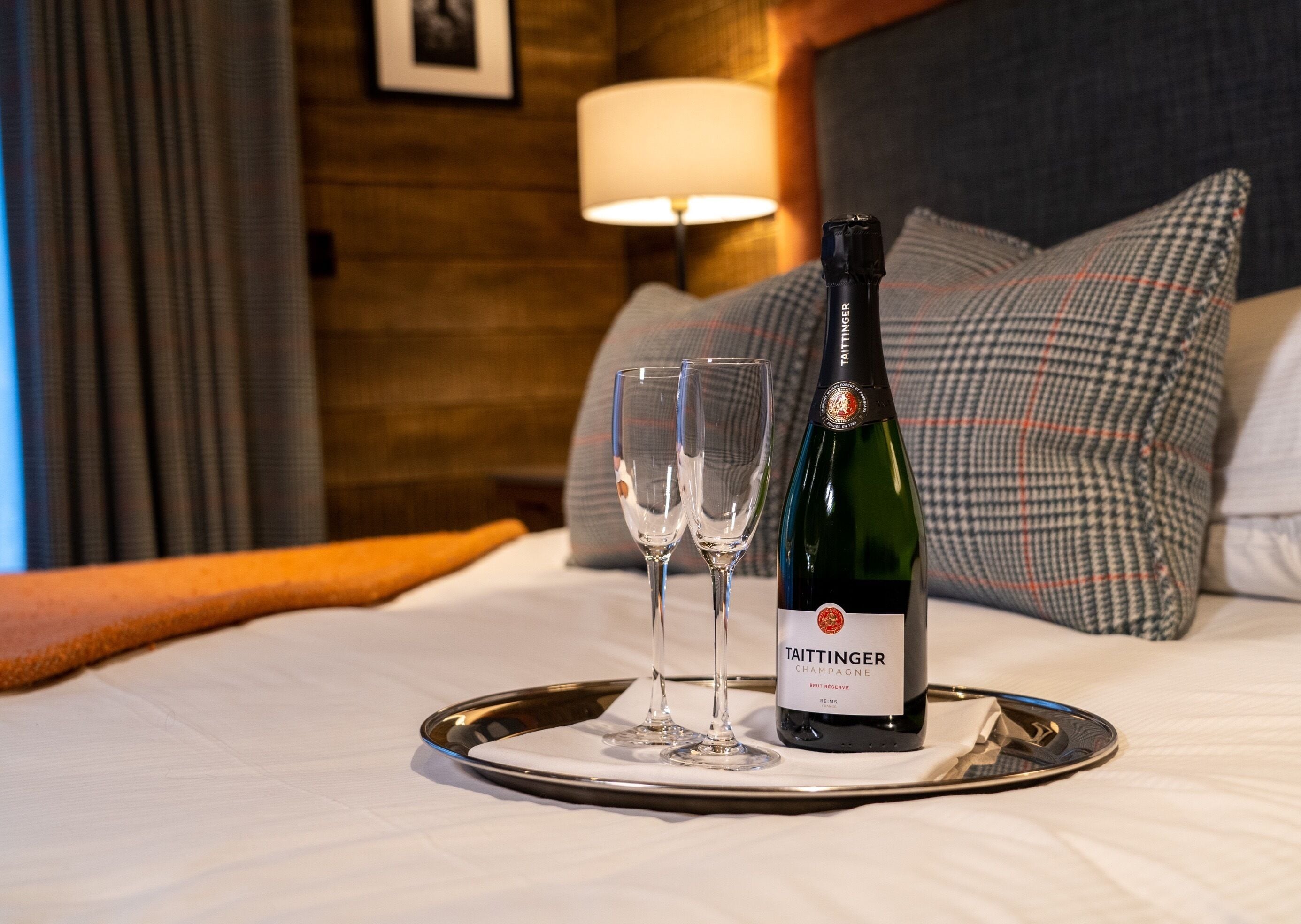 A bottle of Taittinger Champagne on a tray with two glasses on a bed with a headboard and pillows.