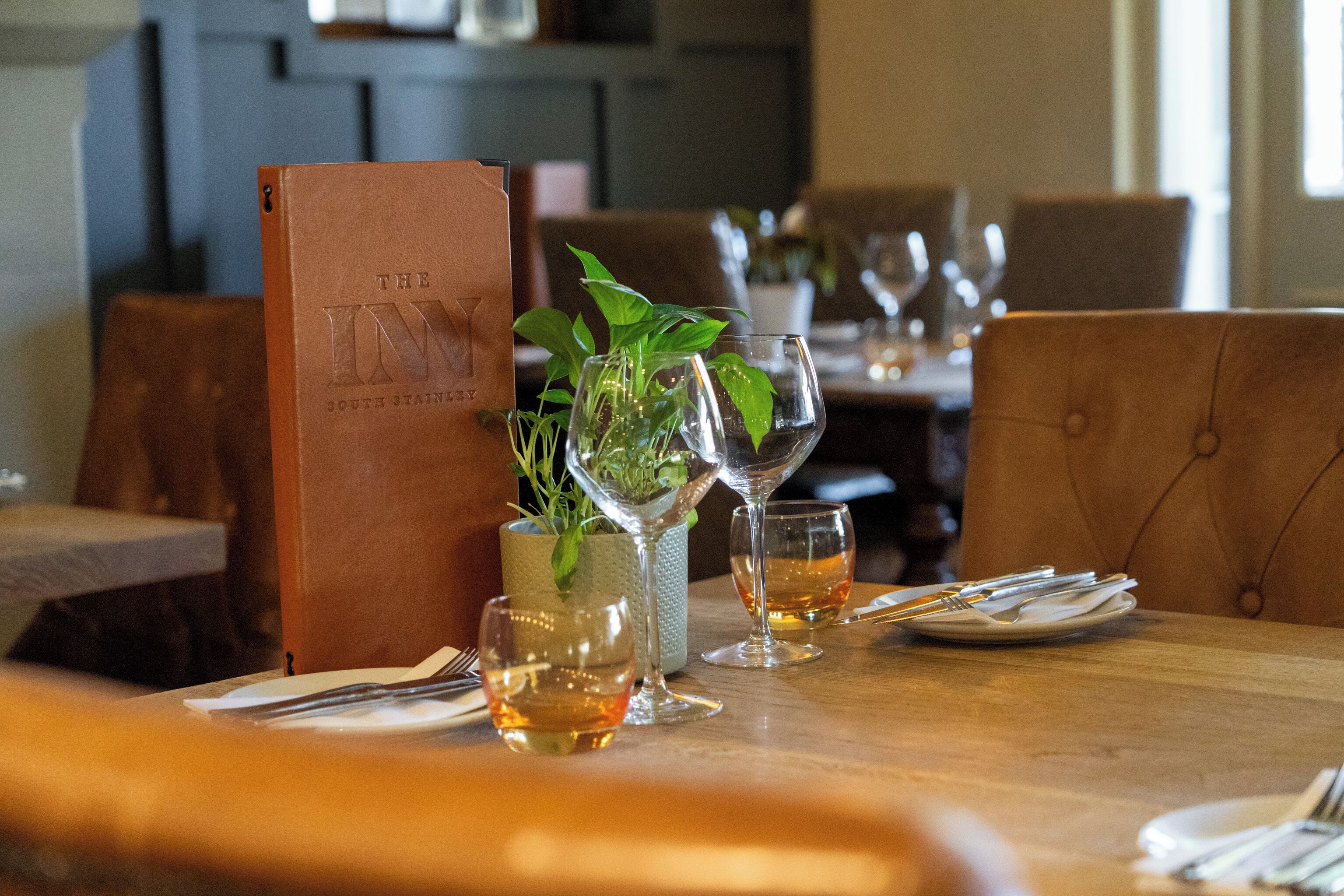 Une table de restaurant mise avec des verres à vin, un menu et une plante en pot.