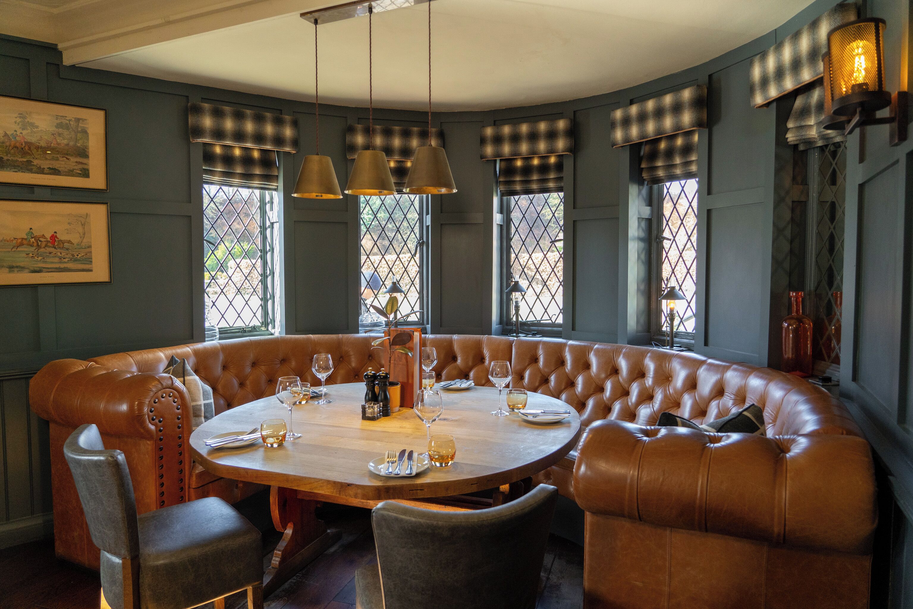 A round table with a leather bench seating, surrounded by chairs, in a room with large windows and checkered window treatments.