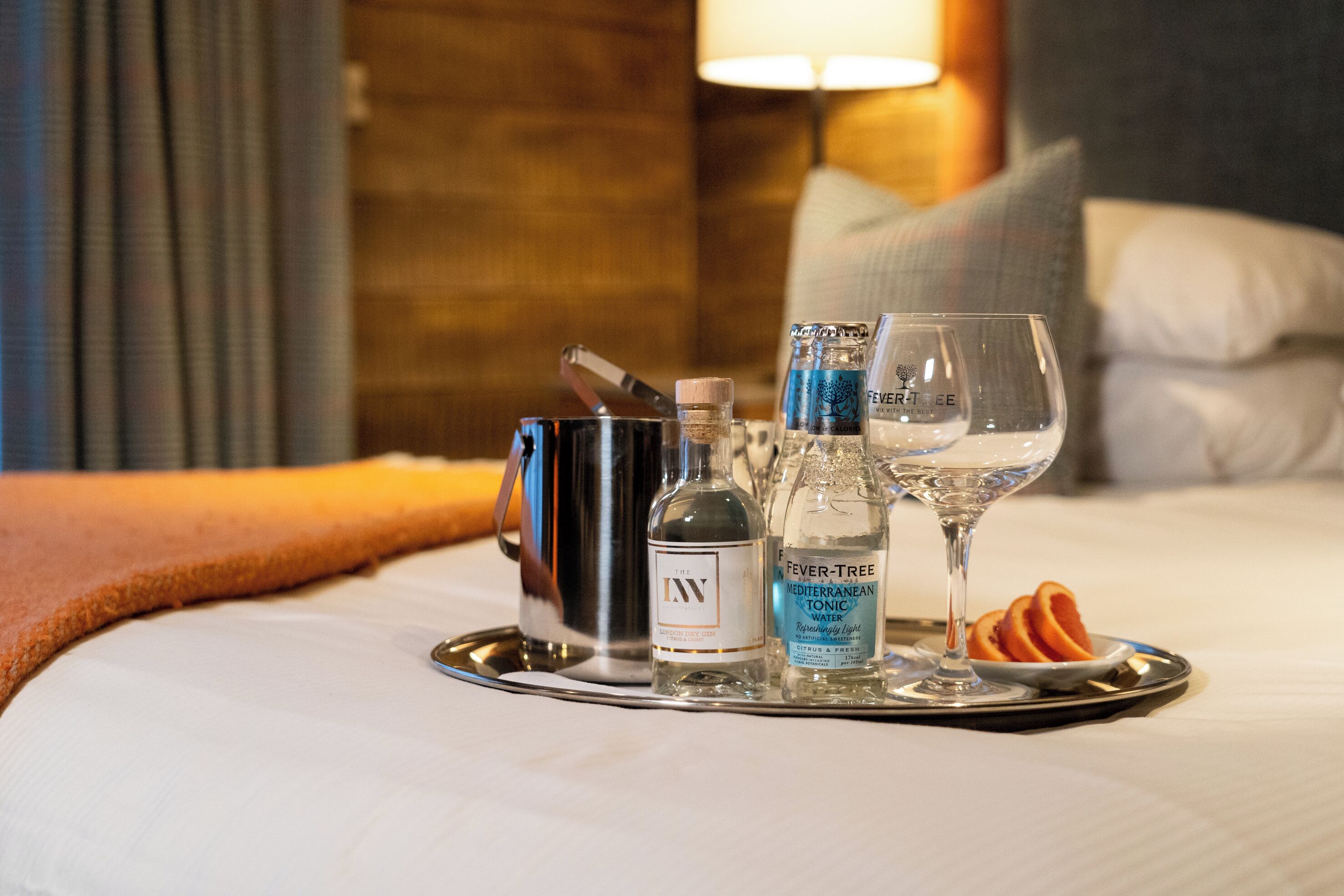 A tray with a bottle of alcohol, a glass, and a small bottle of tonic water on a bed.