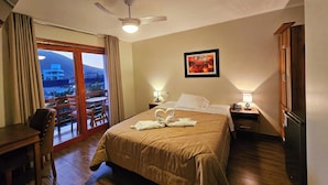 Habitación de hotel con cama, escritorio, silla, mesita de noche, lámpara, ventilador de techo y vistas al exterior.