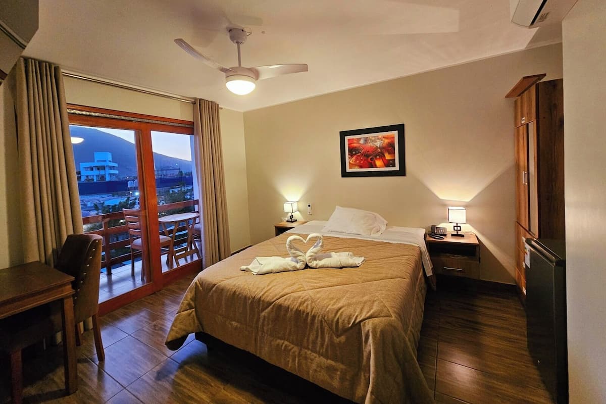 Habitación de hotel con cama, escritorio, silla, mesita de noche, lámpara, ventilador de techo y vistas al exterior.