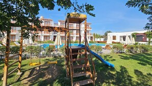 Un parque infantil con tobogán y columpios, rodeado por una piscina y un edificio con balcones.