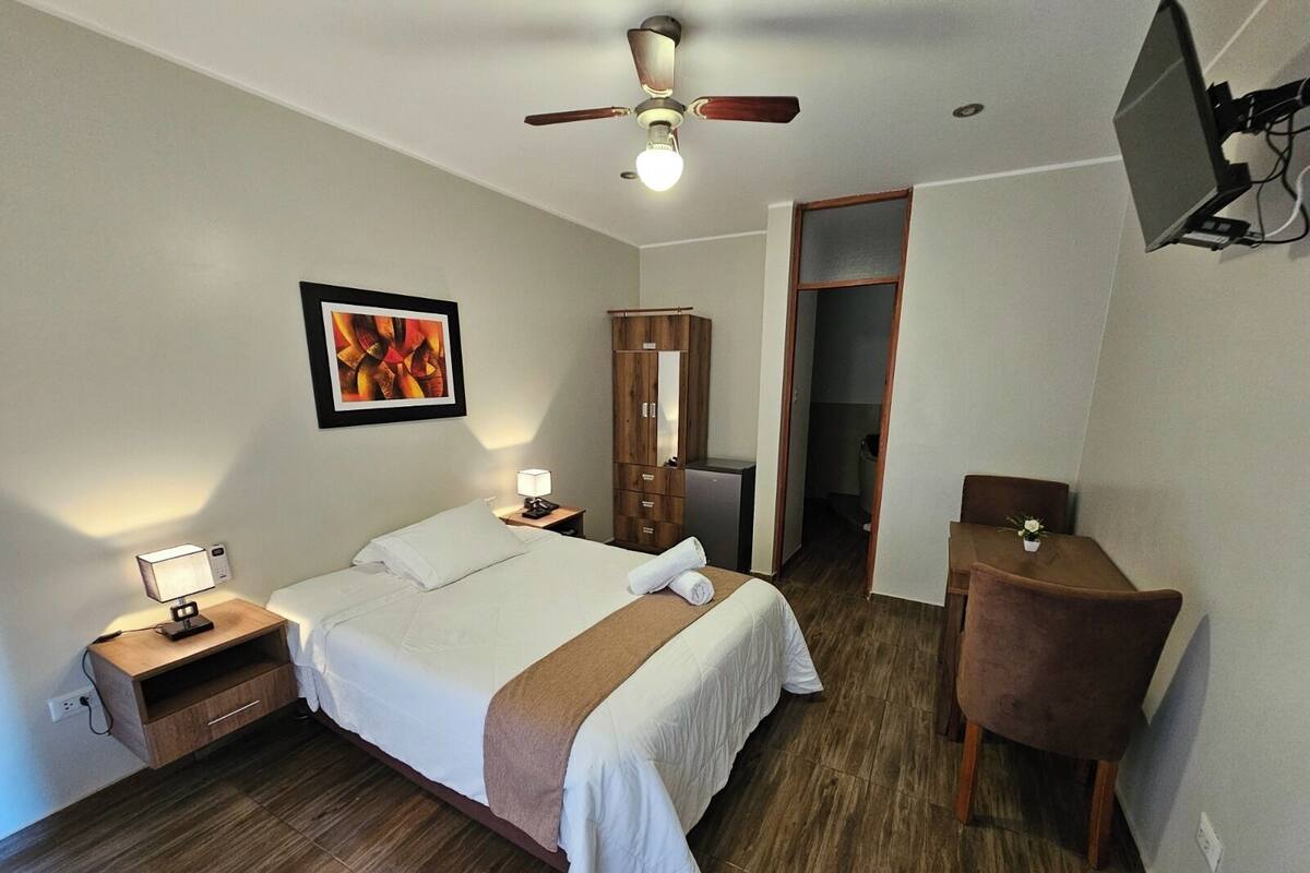 Una habitación de hotel con cama, mesitas de noche, escritorio, silla, ventilador de techo, televisor y puerta que da a un baño.
