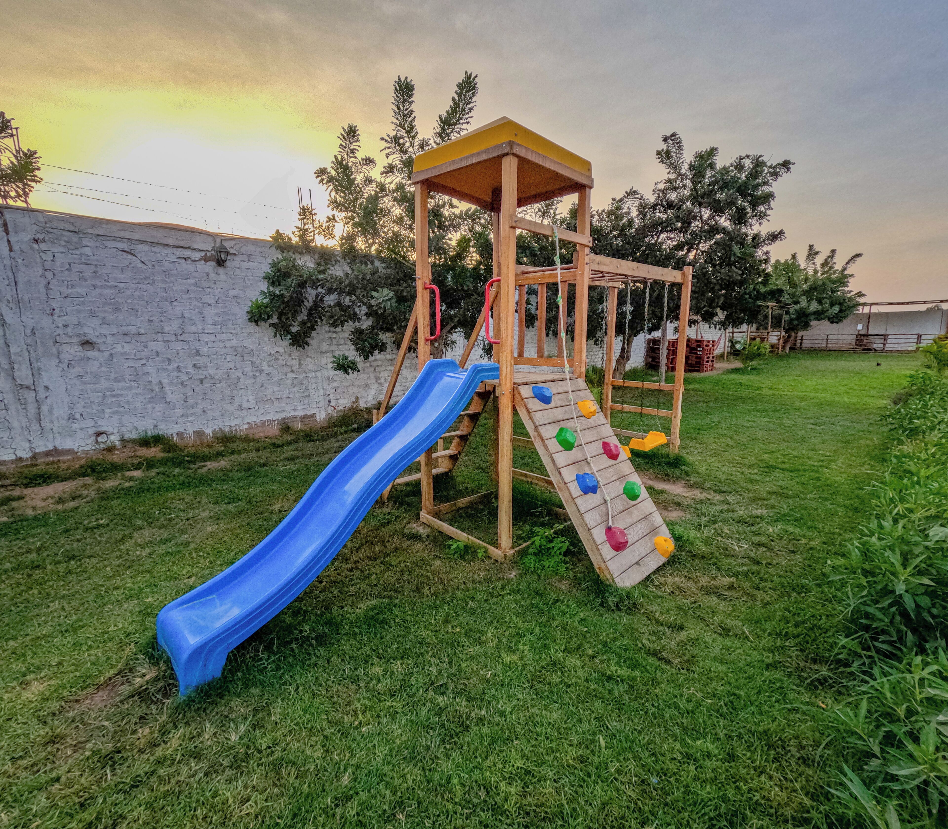 A wooden playground structure with a blue slide and climbing steps, set against a white wall and surrounded by grass and trees.