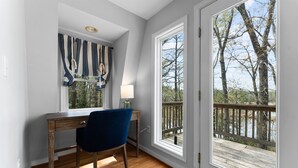 Une pièce avec un bureau en bois, une chaise bleue, une lampe et de grandes fenêtres donnant sur des arbres et une terrasse.