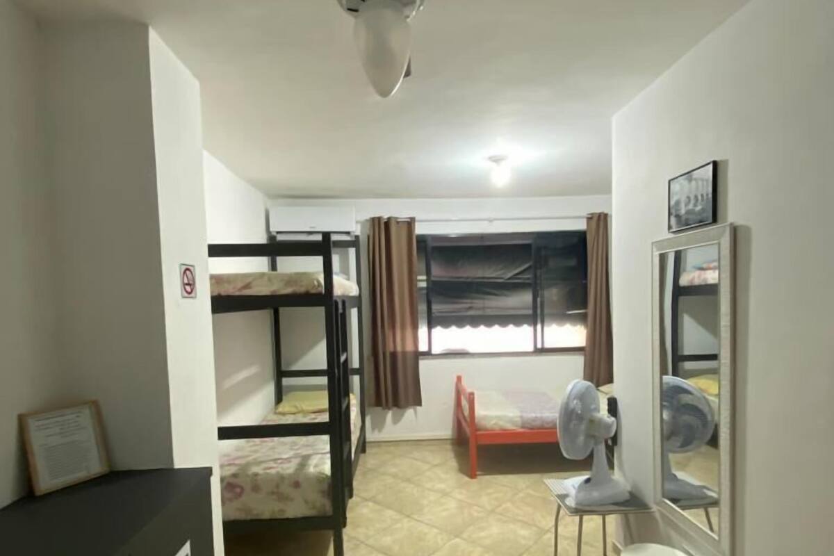 A dormitory room with bunk beds, a ceiling fan, a window with curtains, a desk, and a cabinet.