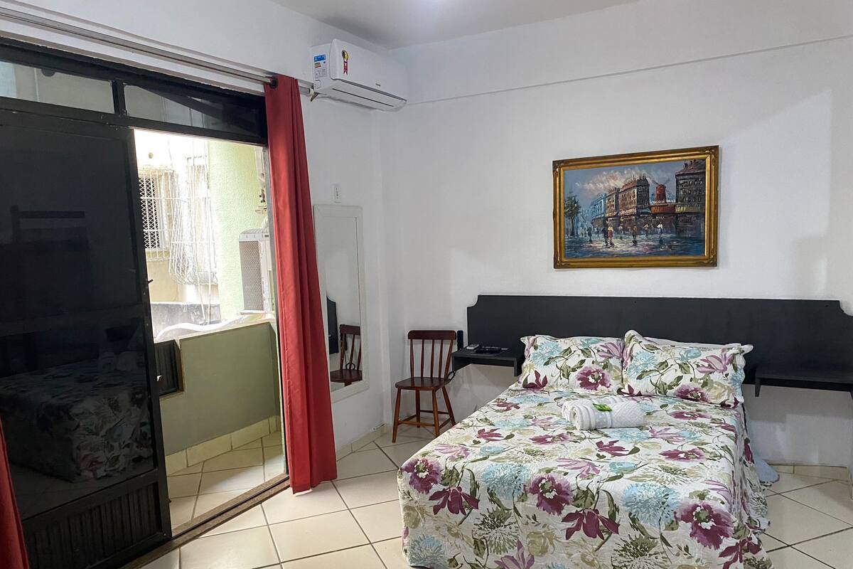 A bedroom with a bed, a chair, a desk, a painting, and an air conditioning unit.