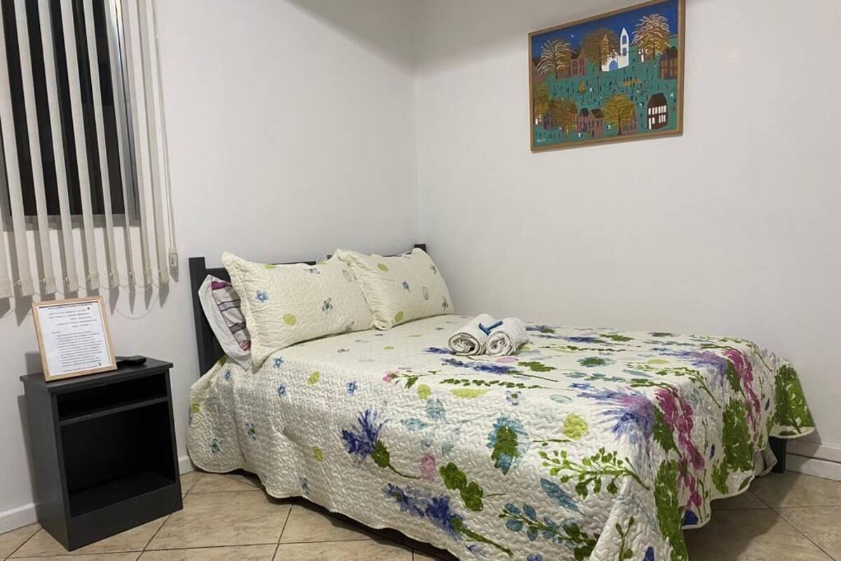 A bedroom with a bed, a nightstand, a wall-mounted painting, and a window with blinds.