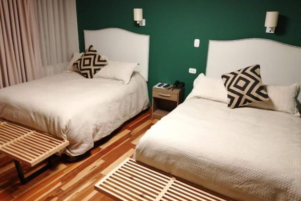 Two beds with white bedding and geometric-patterned pillows, a wooden bench, and a green wall with two wall-mounted lights.